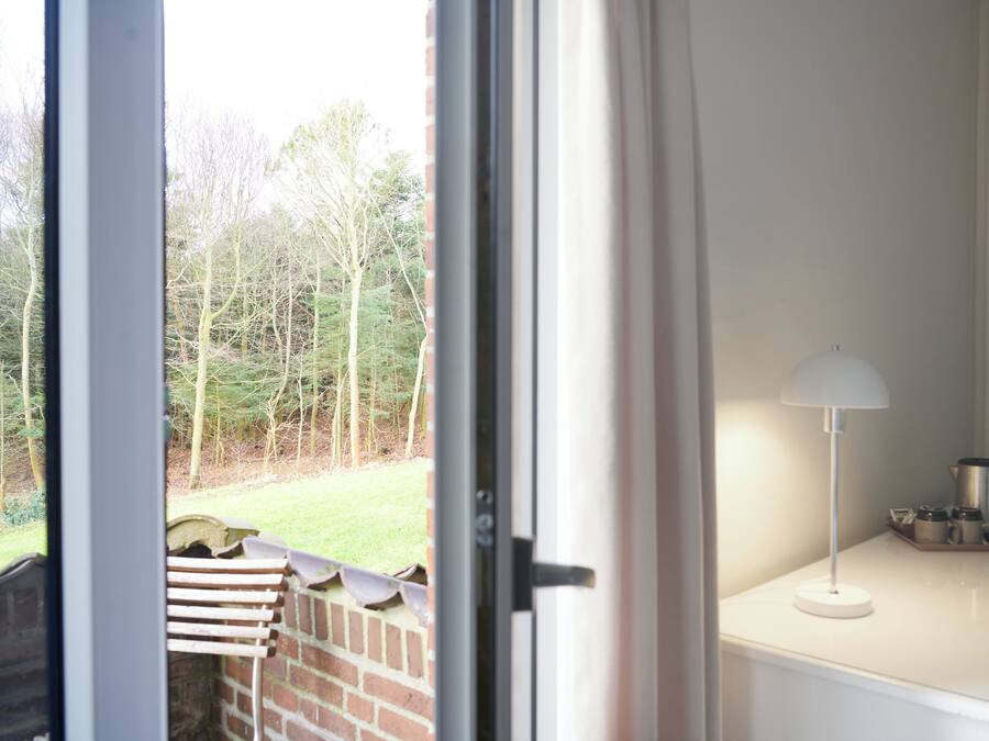 A room with a white lamp, a coffee maker, and a view of a garden through an open door.
