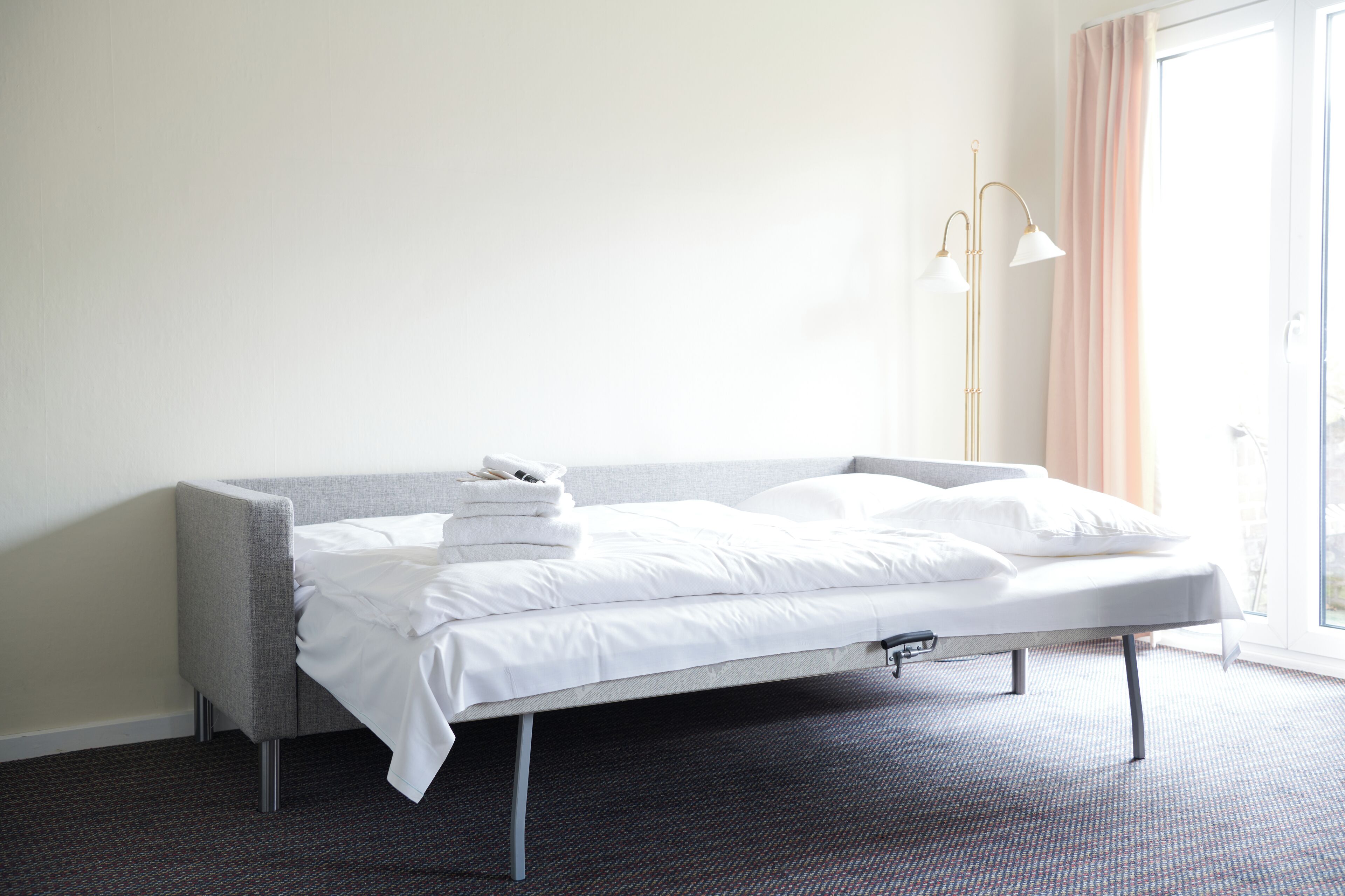 A neatly made bed with white linens, a grey upholstered headboard, and a modern floor lamp.