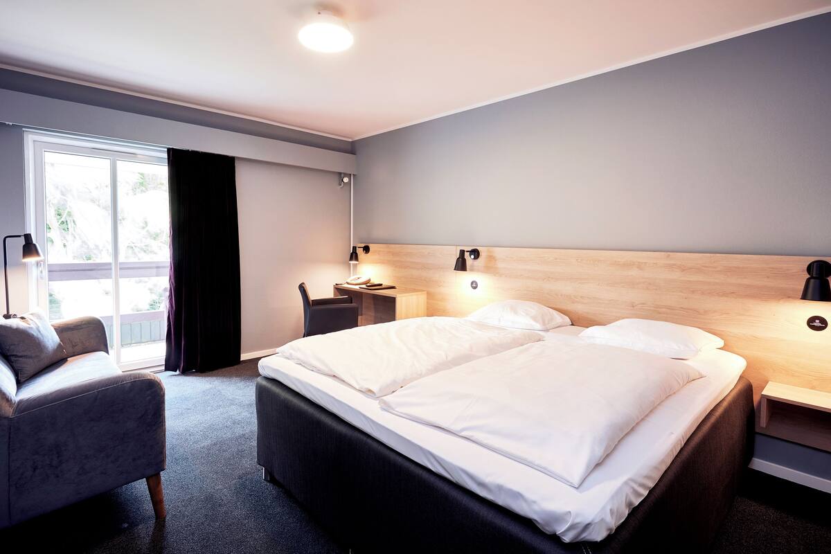 Una habitación de hotel moderna con una cama grande, un escritorio, una silla y un ventanal con cortinas.