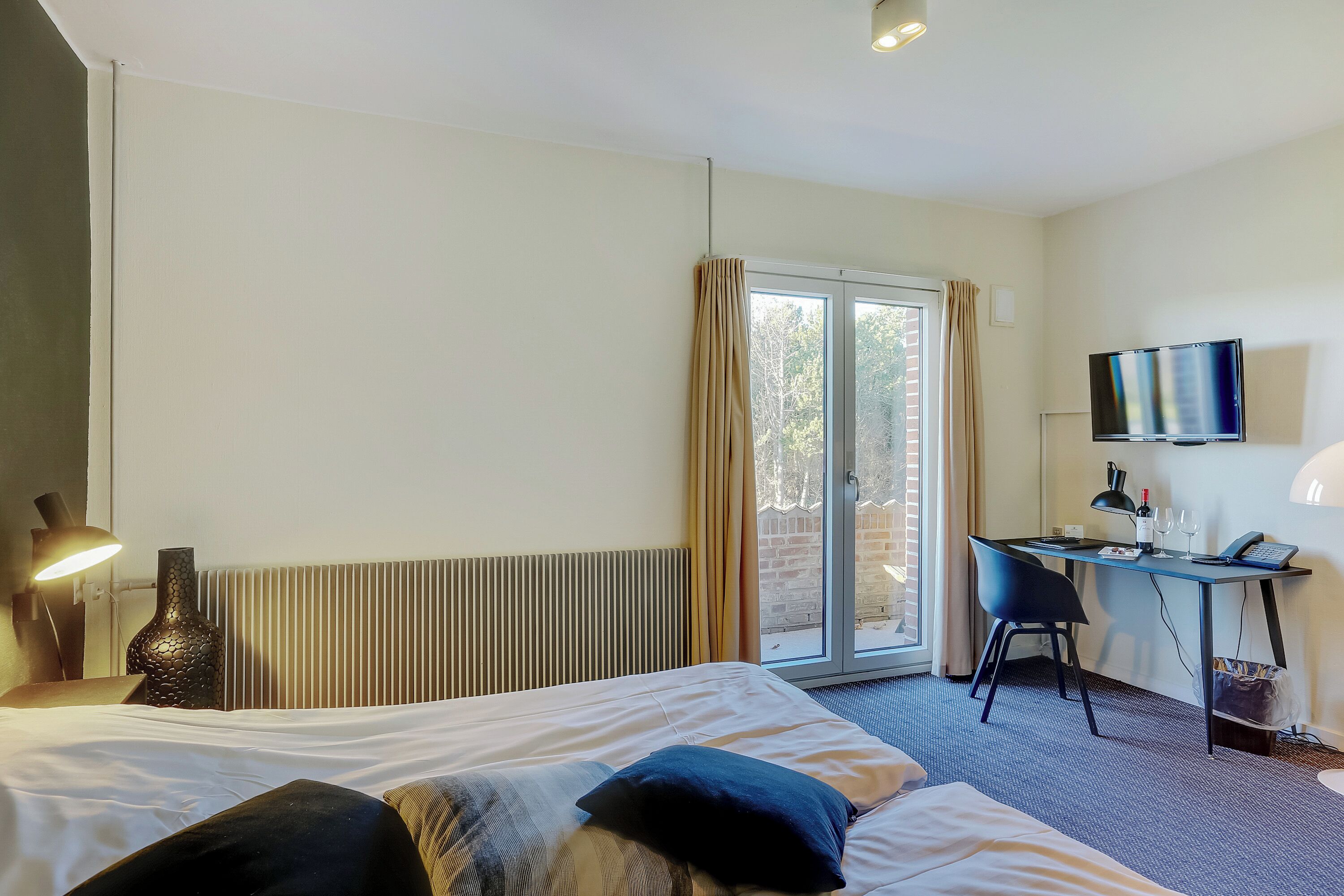 A hotel room with a bed, a desk with a chair, a TV, and a window with curtains.