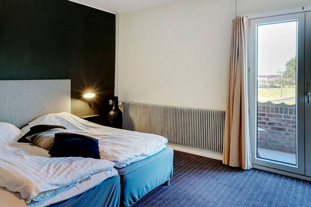 Habitación de hotel con cama, mesita de noche, lámpara, radiador y vistas al exterior.