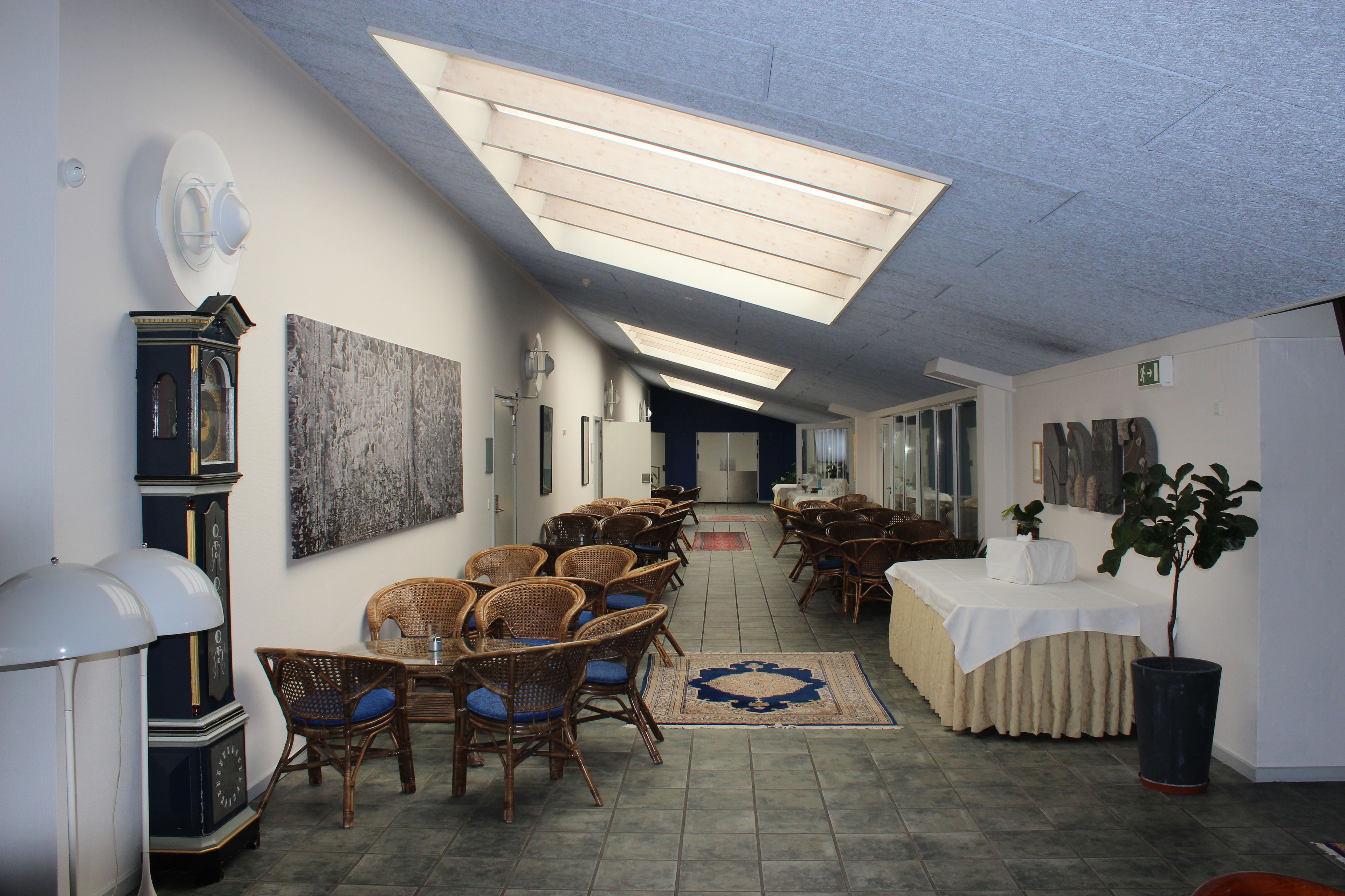A long, narrow room with high ceilings and skylights, furnished with wicker chairs and tables, and decorated with clocks and artwork.