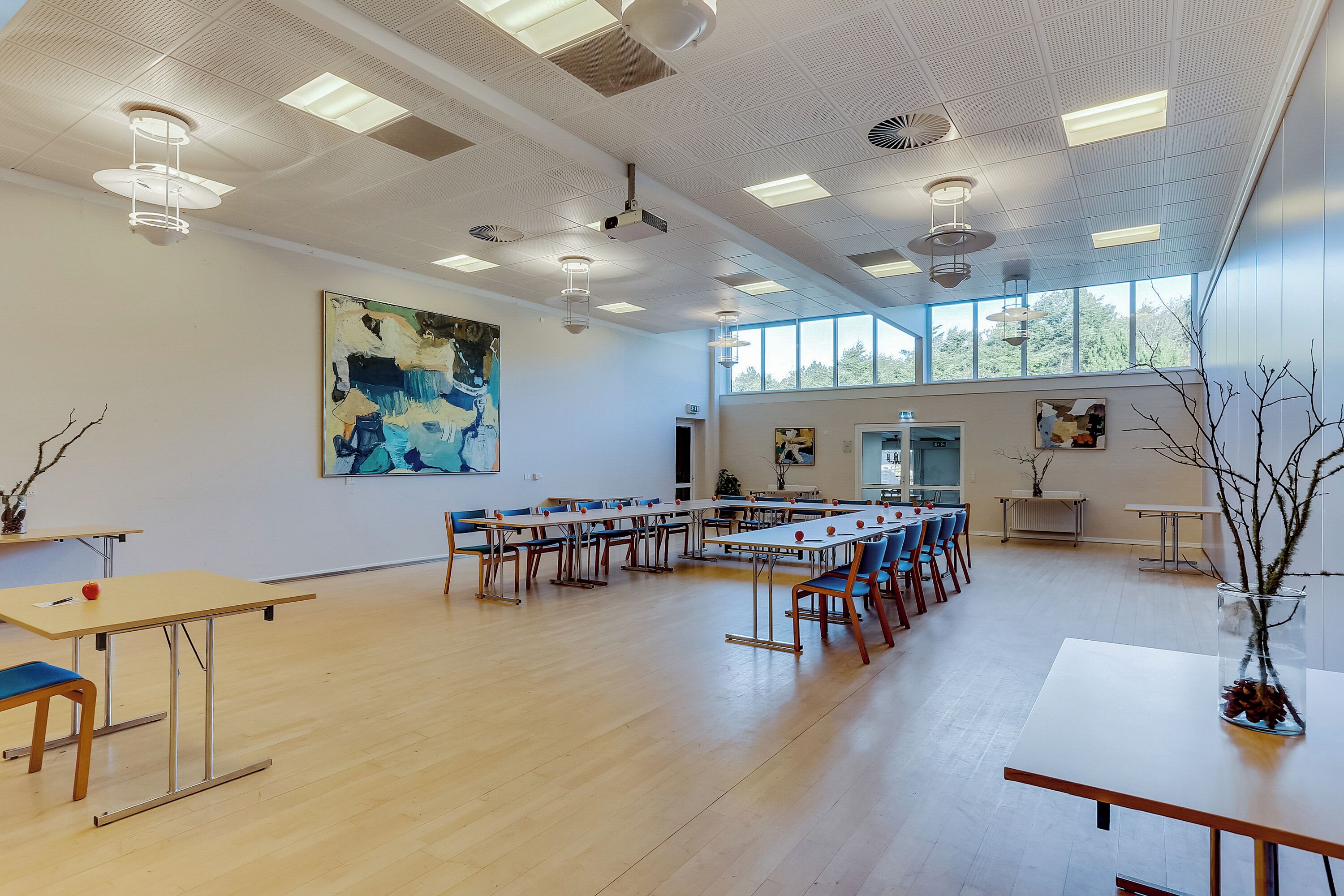 A spacious room with long tables and chairs, ceiling lights, and large windows.