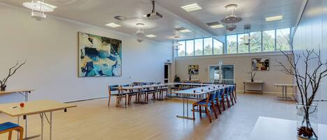 A spacious room with long tables and chairs, ceiling lights, and large windows.