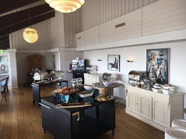 Una habitación amplia con una disposición tipo buffet, que incluye una estación de café, exhibición de alimentos y un área de descanso con un mueble de madera y un cuadro en la pared.