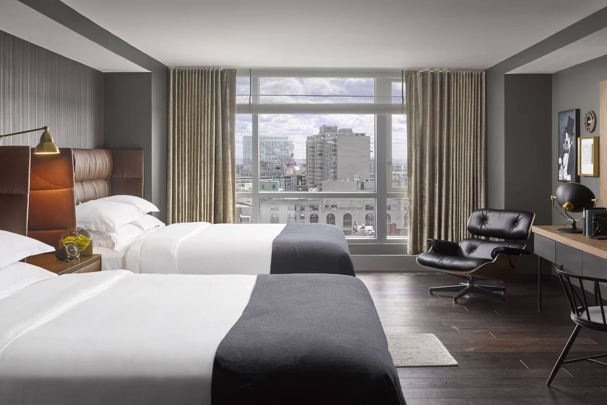 A hotel room with two beds, a desk, a chair, and a large window offering a city view.
