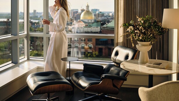 Une chambre d’hôtel moderne avec de grandes fenêtres, une vue sur le paysage urbain, un fauteuil en cuir, une petite table et un vase rempli de fleurs.