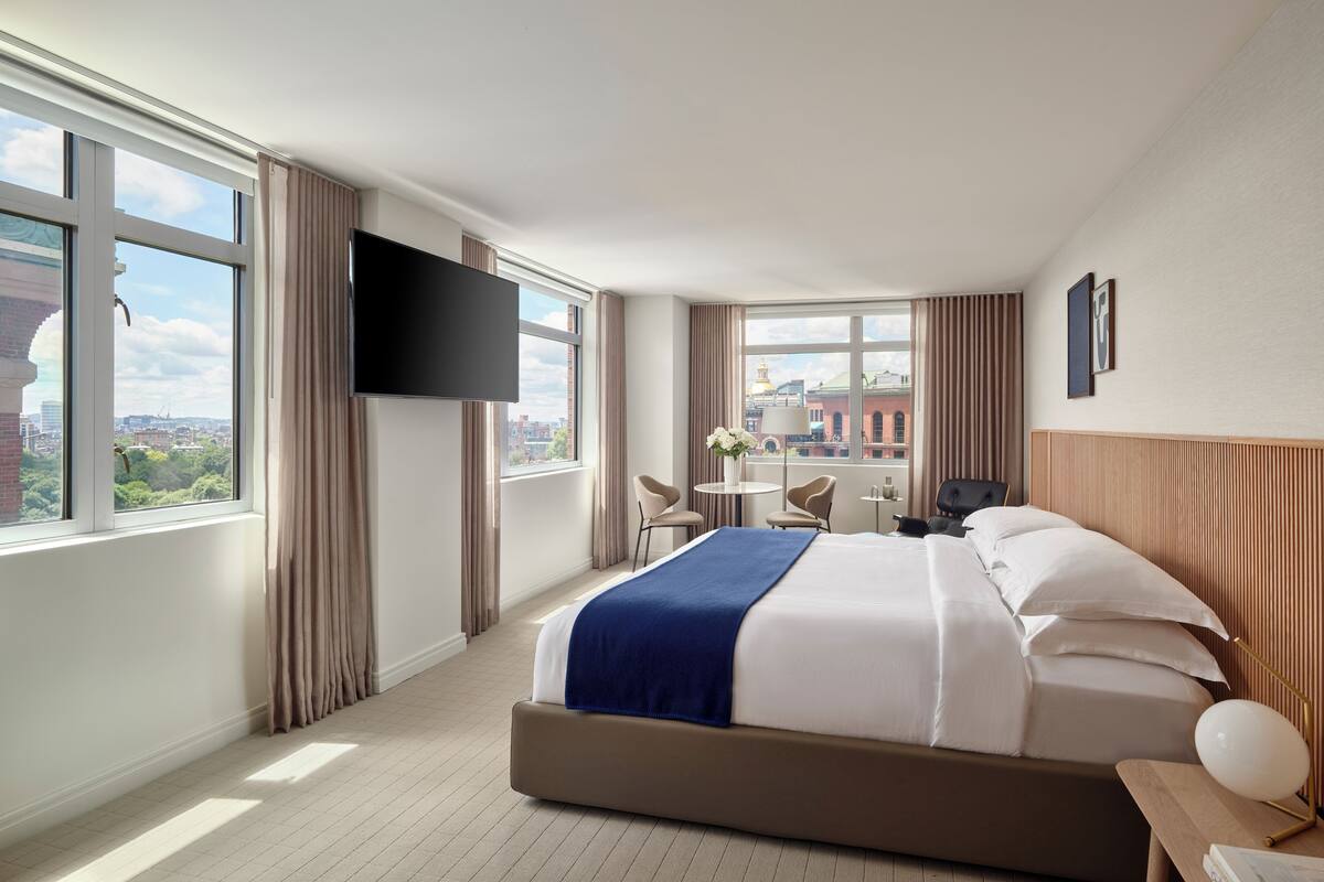 A hotel room with a large bed, a flat-screen TV, a small table with chairs, and a view of a cityscape through large windows.