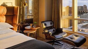 Une chambre d’hôtel avec un grand lit, un fauteuil en cuir, une petite table ronde avec une horloge, et une vue sur la ville grâce à de grandes fenêtres.