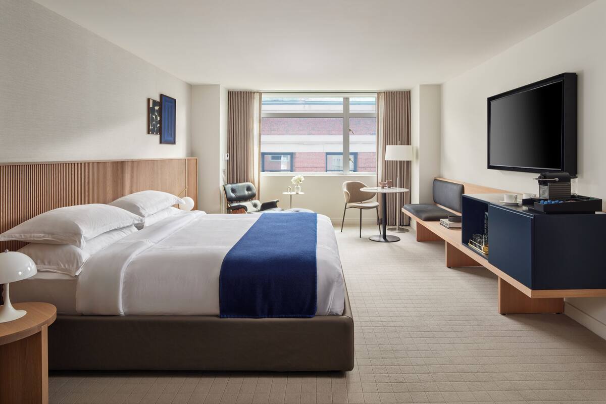 A modern hotel room with a large bed, a desk, a chair, a TV, and a window with a view of the city.