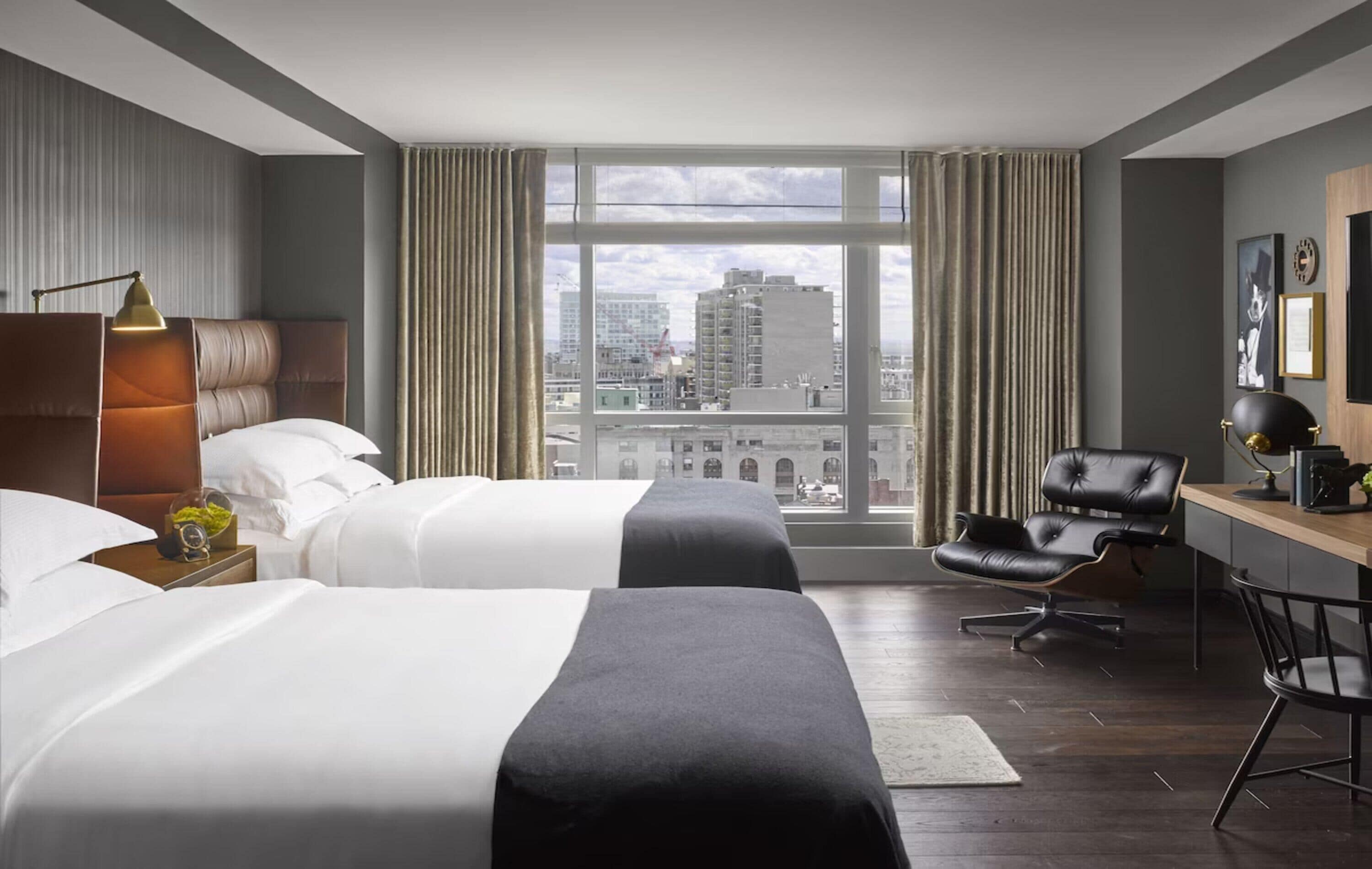 A hotel room with two beds, a desk, a chair, a lamp, and a large window with a city view.