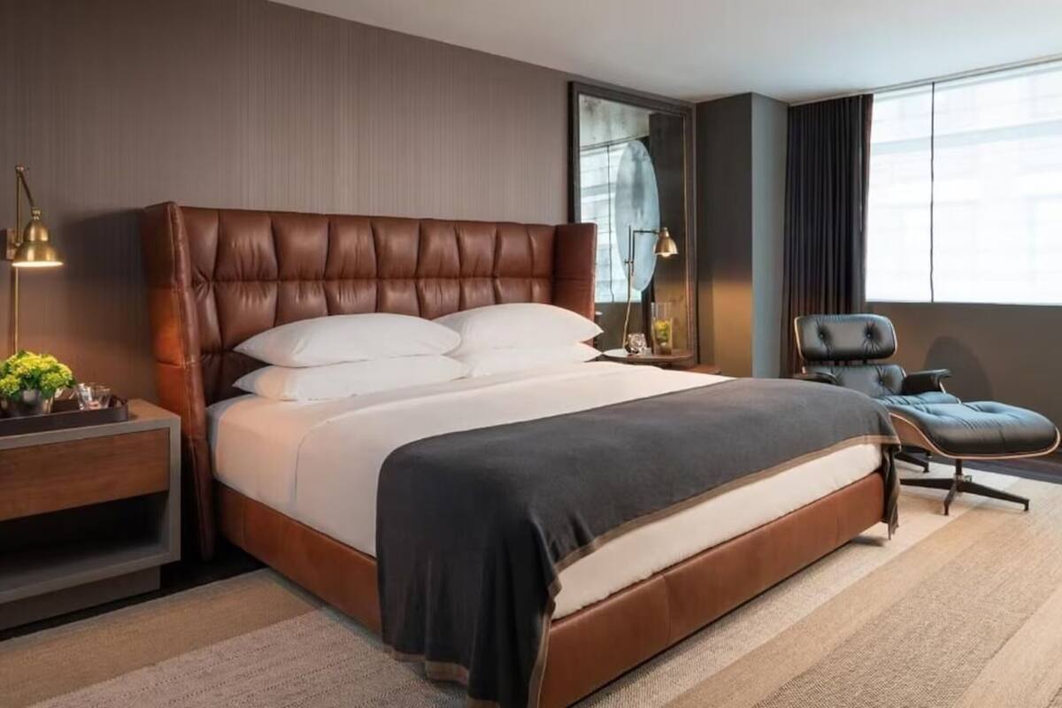 A modern hotel room with a large bed, a nightstand, a chair, and a large window.