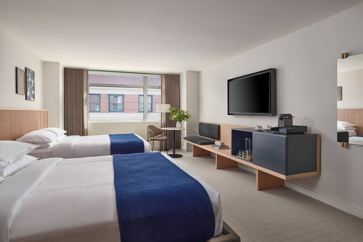 A hotel room with two beds, a desk, a TV, and a window with a view of buildings.