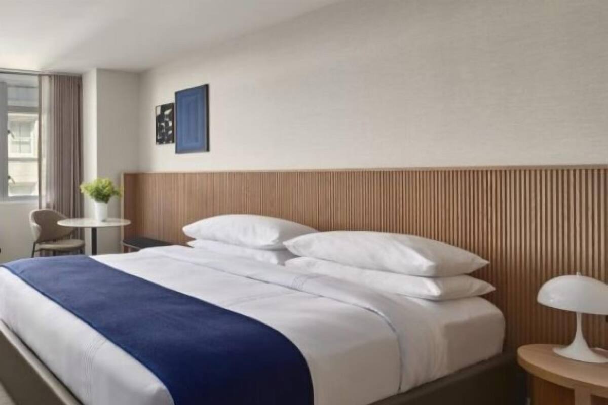 A hotel room with a bed, a bedside table with a lamp, a small table with a plant, and a window with curtains.