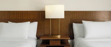 A hotel room with two beds, a wooden headboard, a nightstand with a lamp, and a small drawer.