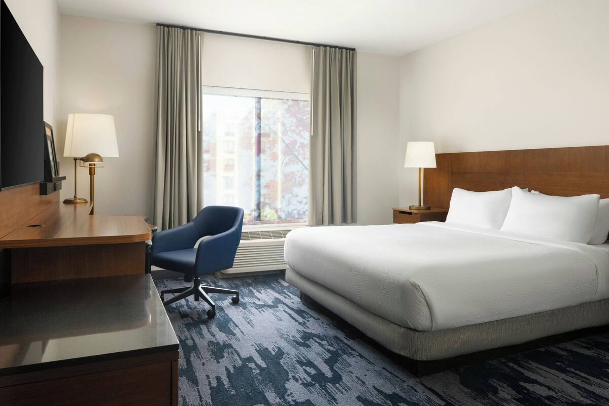 Une chambre d’hôtel avec un lit, un bureau, une chaise, une lampe et une fenêtre avec des rideaux.