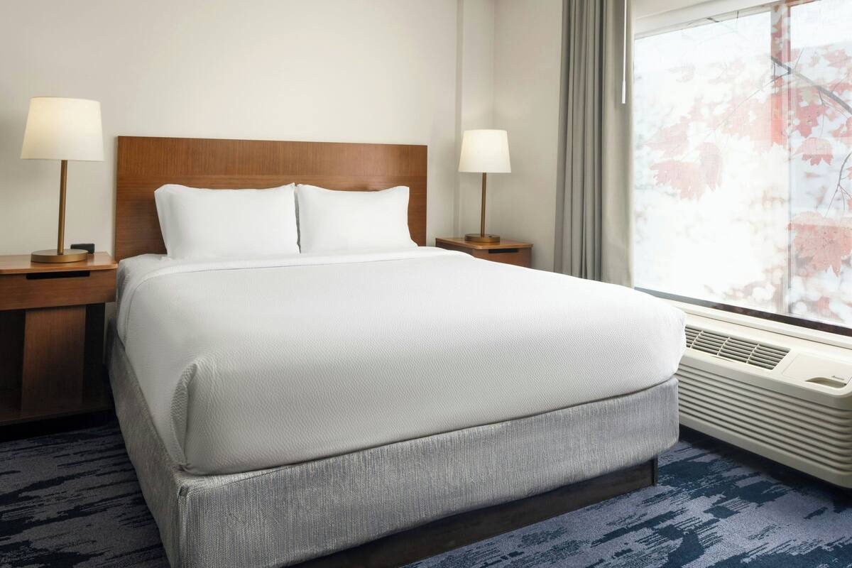 Une chambre d’hôtel avec un grand lit, deux tables de chevet avec des lampes, une fenêtre donnant sur des arbres et un système de climatisation intégré.