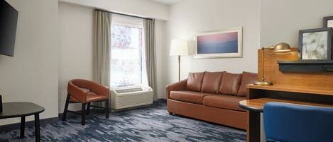 A hotel room with a brown leather sofa, a blue chair, a small table, a lamp, and a painting on the wall.