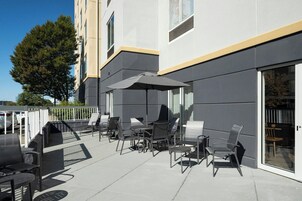 Un patio al aire libre con sillas y mesas modernas, un paraguas grande y un edificio con puerta de cristal.
