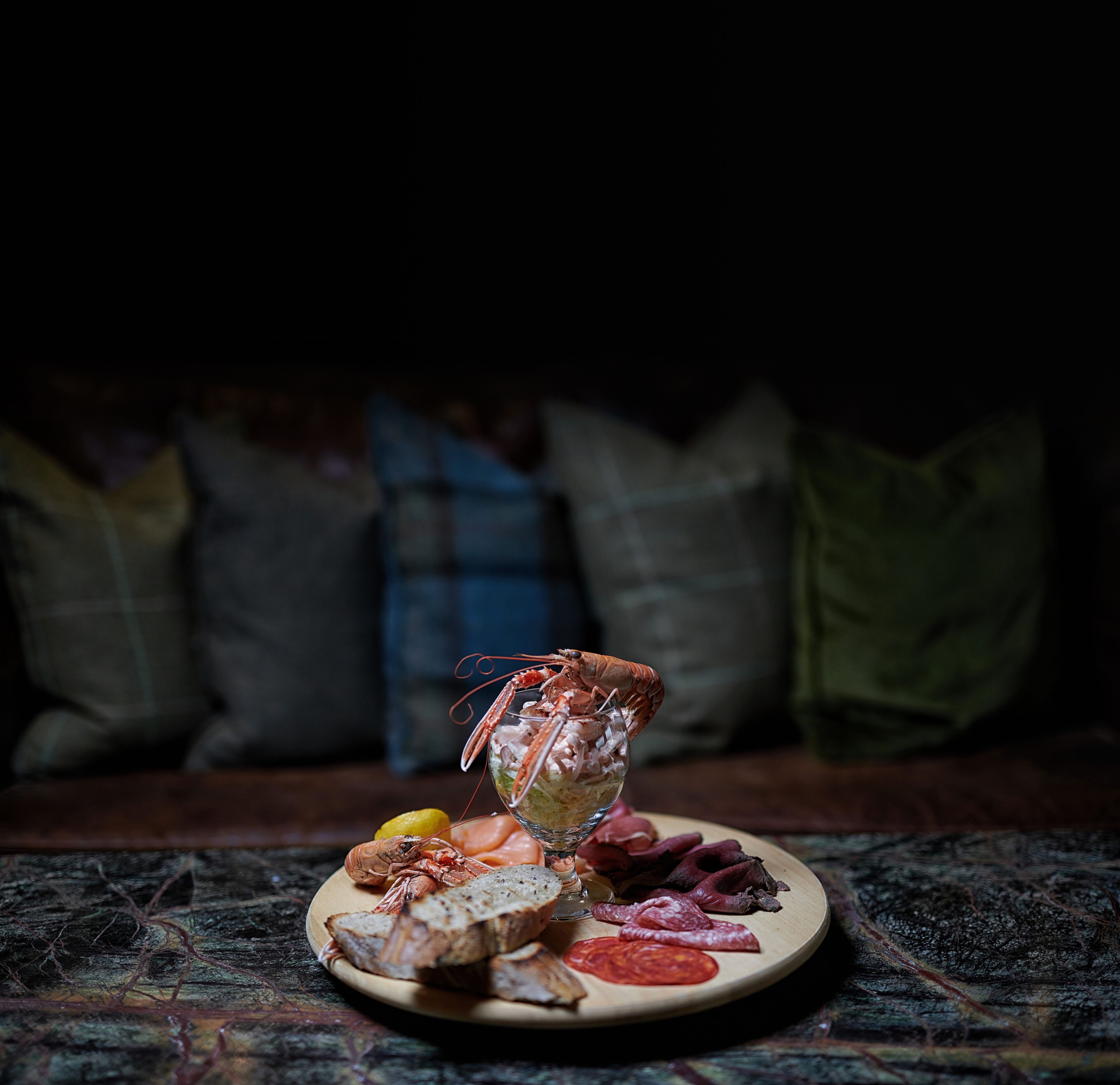 A seafood platter with a lobster on a plate, accompanied by bread, tomatoes, and various meats, set on a table with a dark background and pillows.