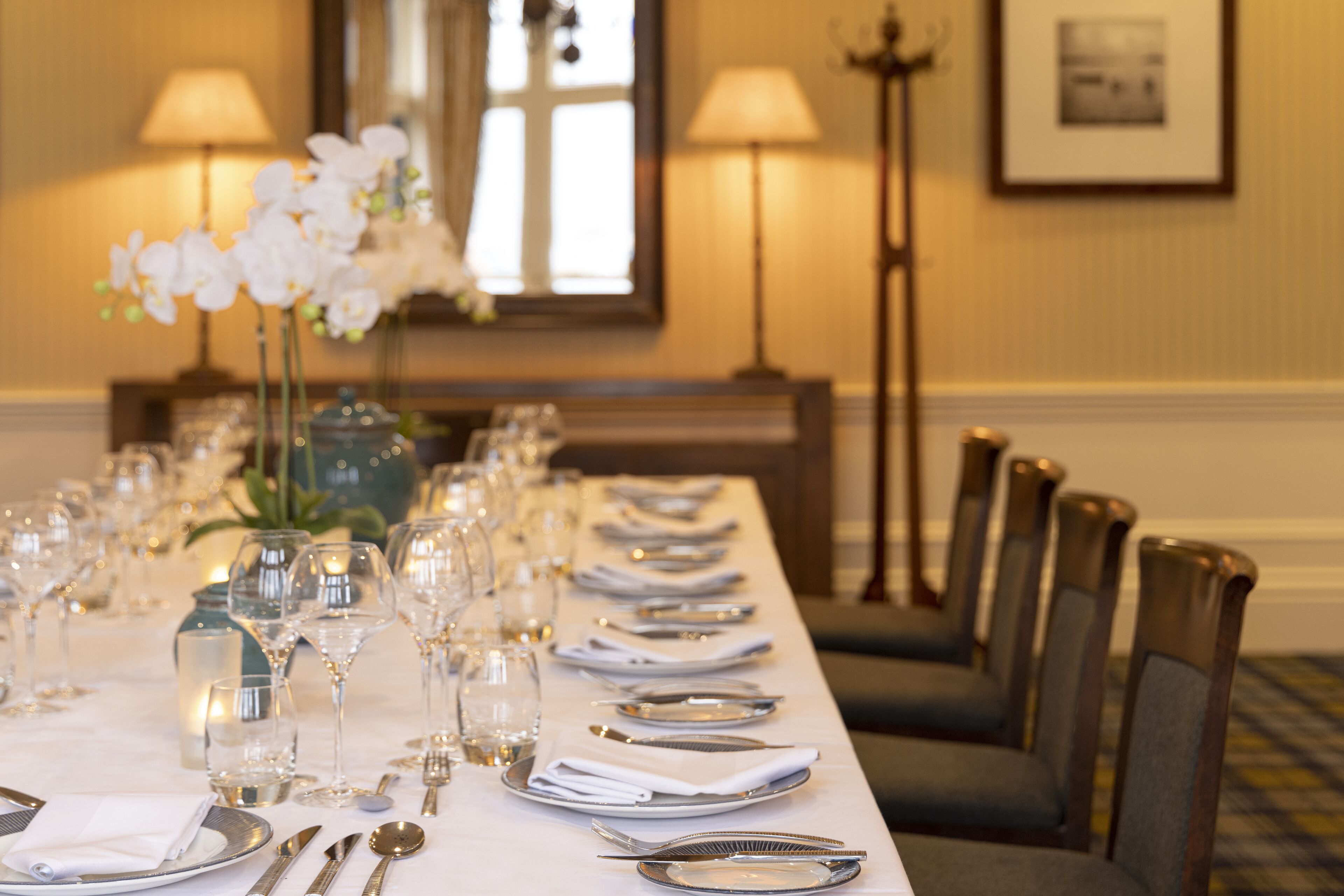 A dining table set with multiple plates, cutlery, and glasses, flanked by chairs and lamps.