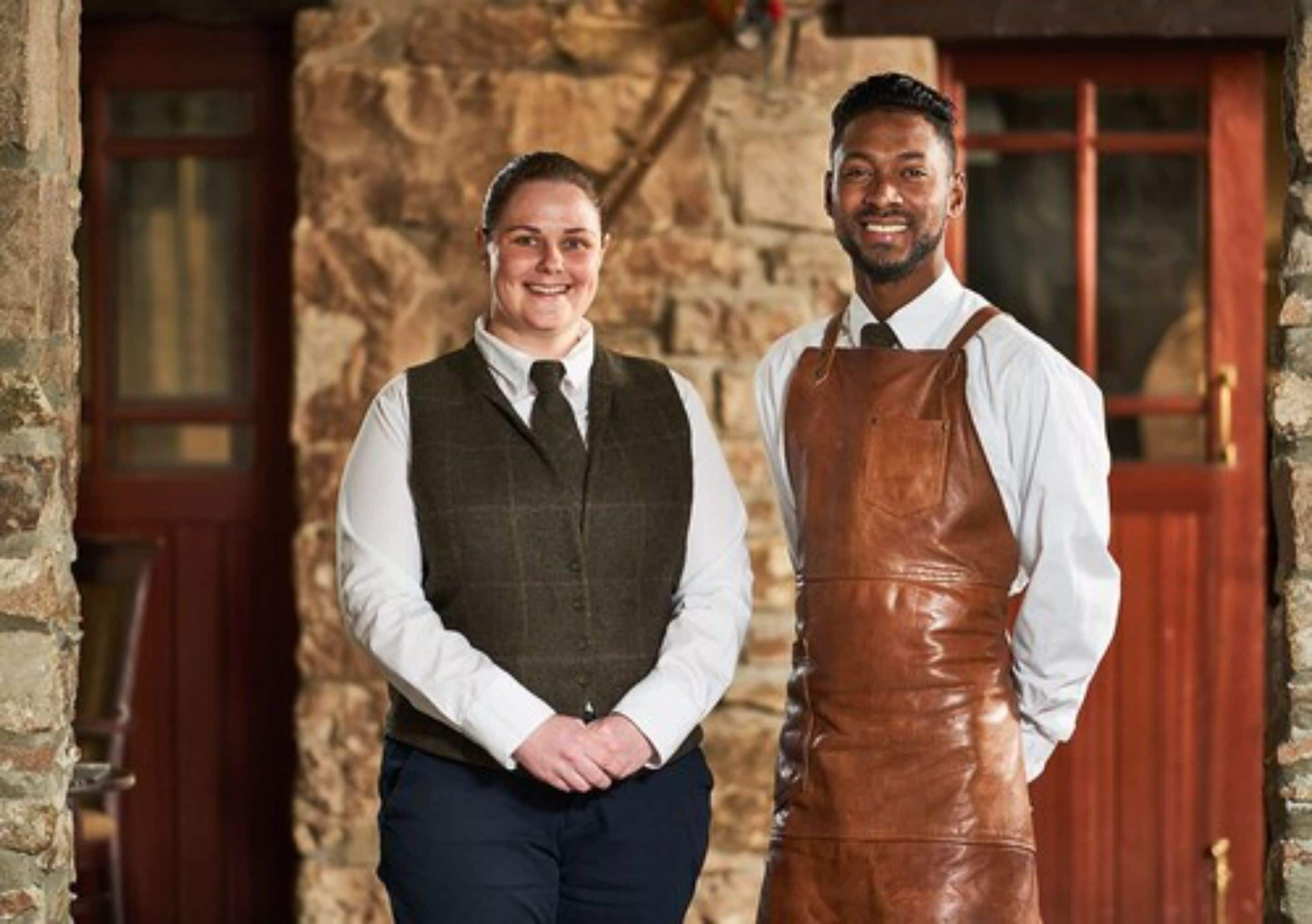 Two individuals standing in front of a stone building with wooden doors.