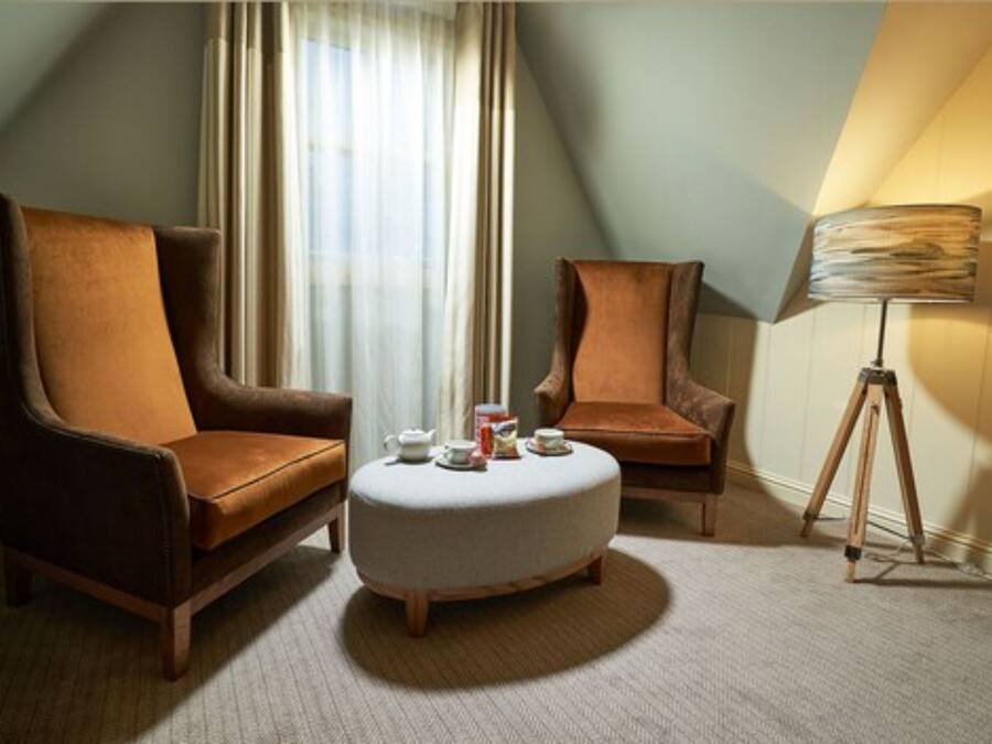 A hotel room with two brown armchairs, a white ottoman, a lamp, and a window with curtains.