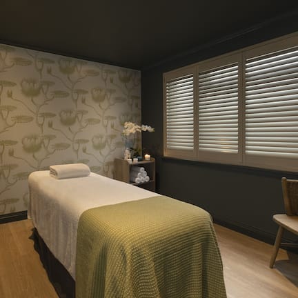 A spa room with a massage table, a chair, a vanity with a mirror, and a window with blinds.