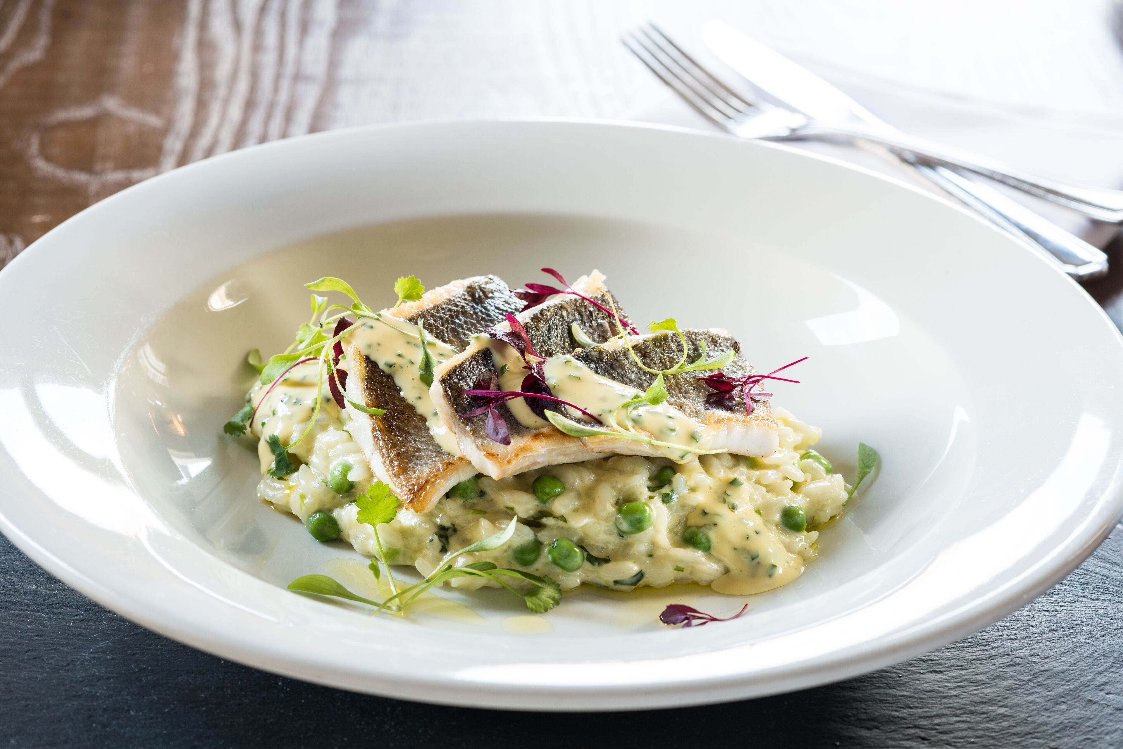 A white plate with a fish dish, peas, and greens on a dark surface.