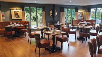 A restaurant with multiple dining tables and booths, large windows with curtains, and a fireplace.