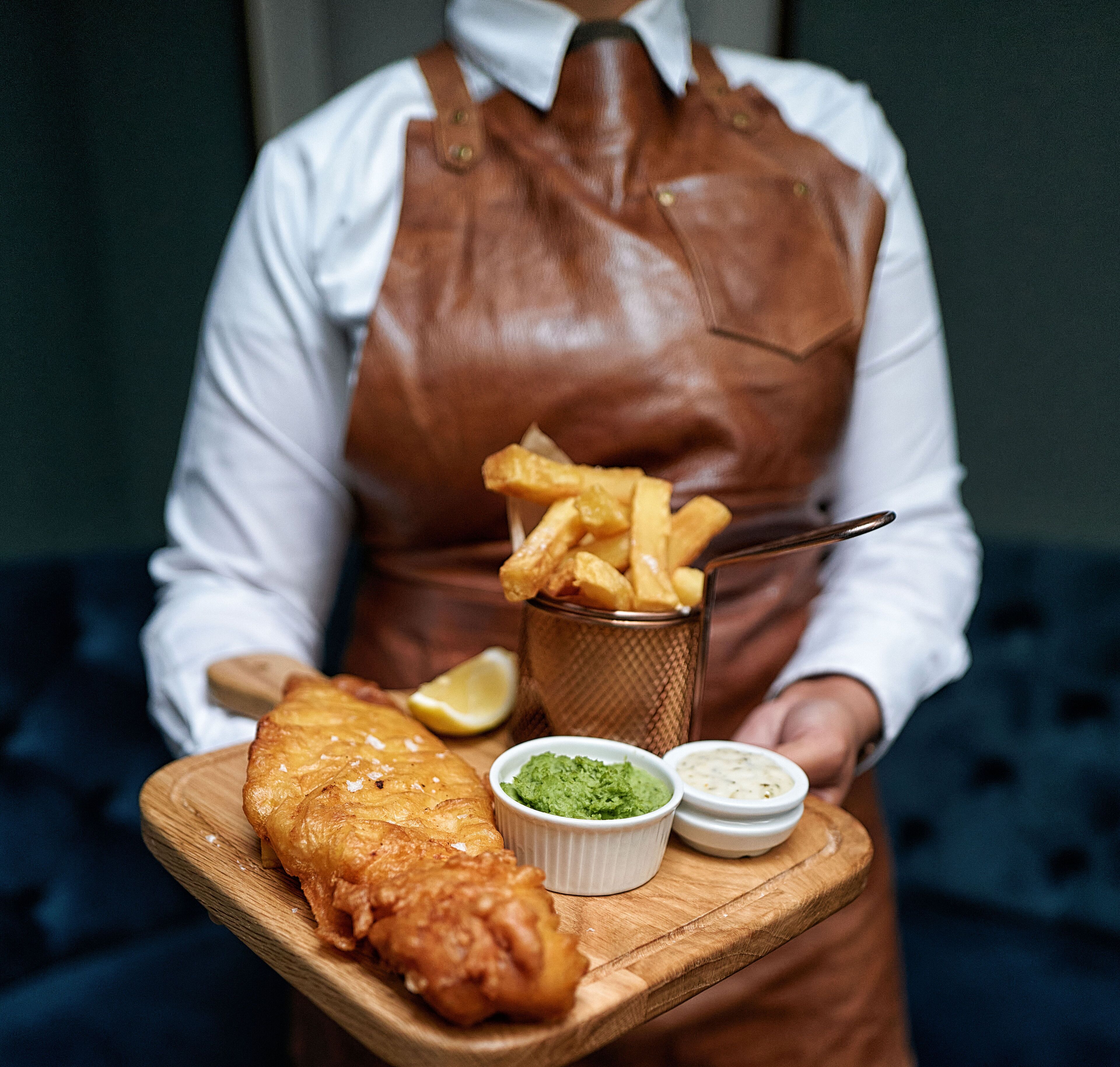 A person serving a fish and chips meal with lemon and two dipping sauces on a wooden board.