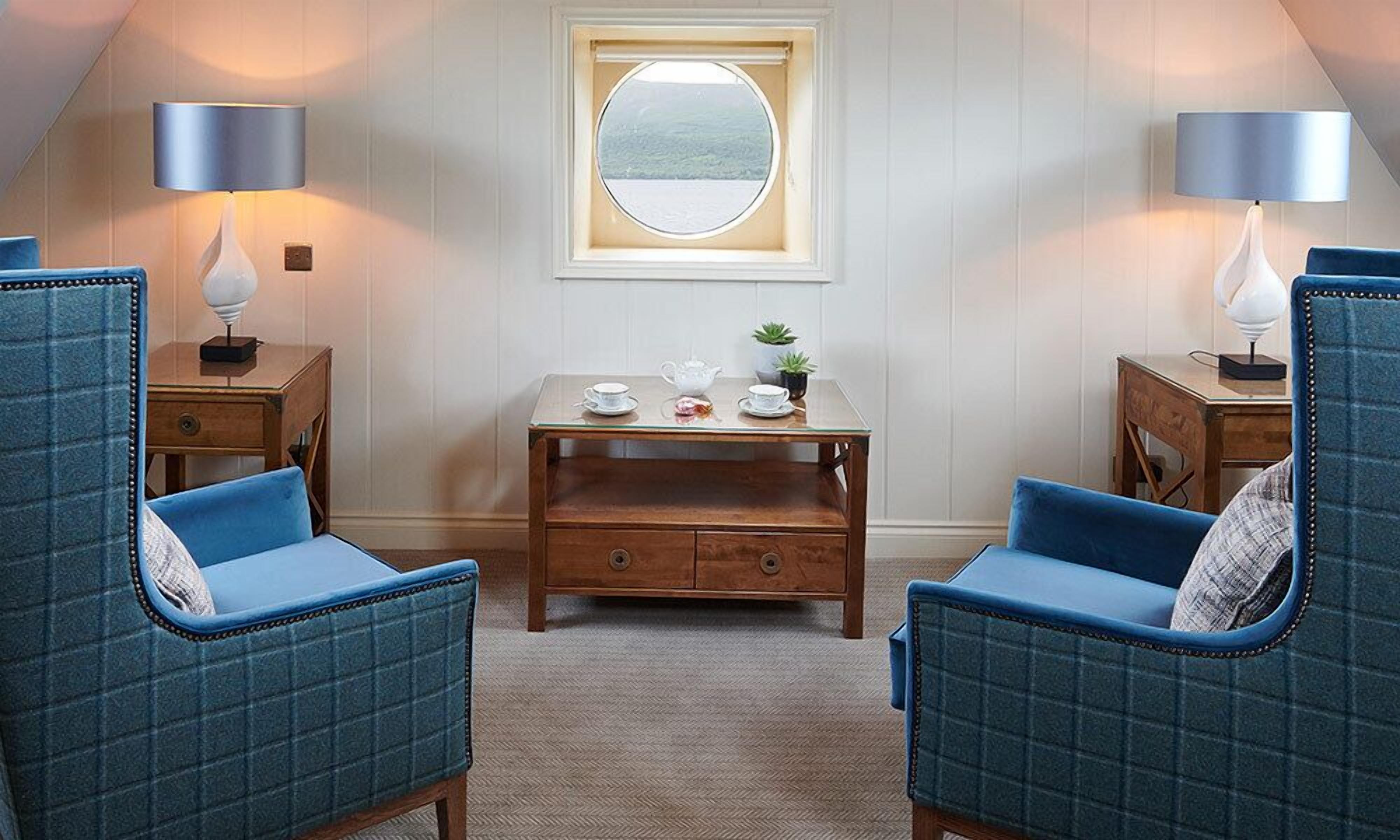A room with two blue upholstered chairs, a small wooden table with a teapot, cups, and a plant, and a window with a view of the outdoors.