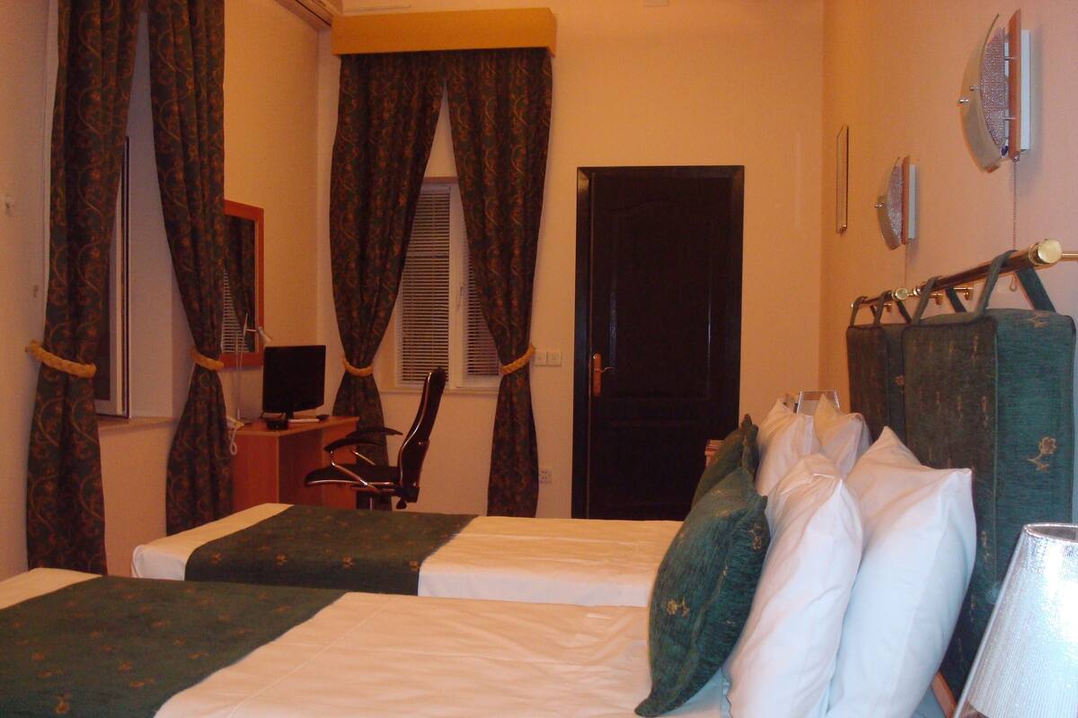 Een hotelkamer met twee enkele bedden, een bureau met een computer, een stoel en een raam met gordijnen.