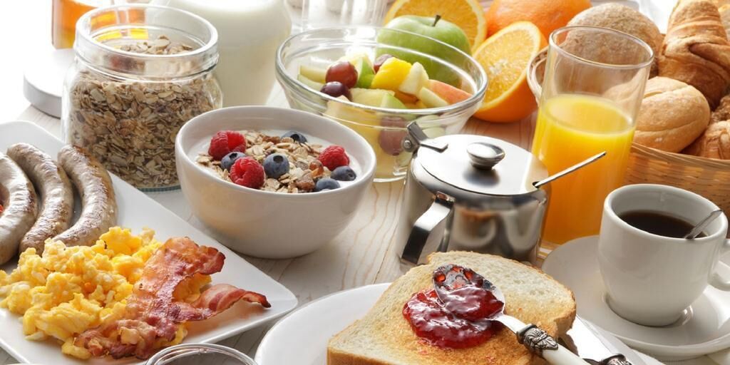 A breakfast table with various food items including sausages, scrambled eggs, toast with jam, cereal with fruits, and orange juice.