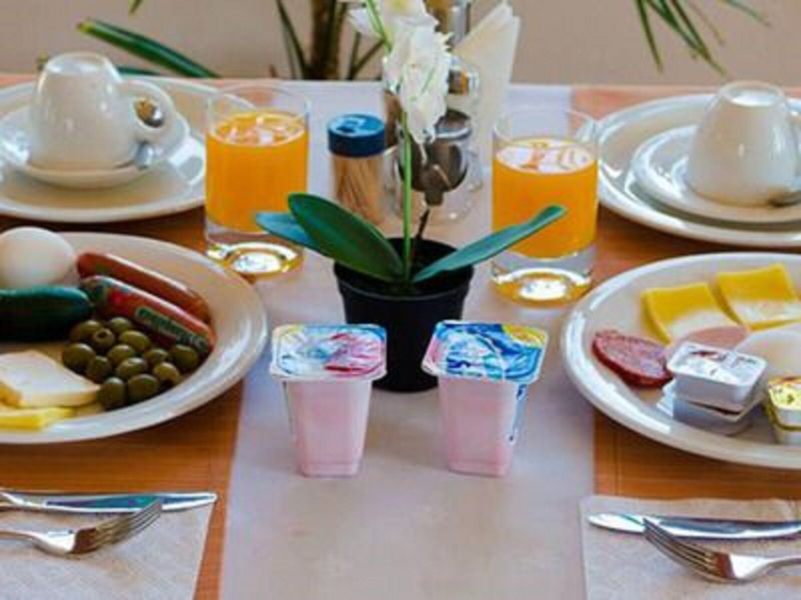 Une table dressée pour le petit-déjeuner, avec des assiettes contenant des œufs, des saucisses, du fromage et des olives, accompagnées de tasses de café et de verres de jus d’orange.
