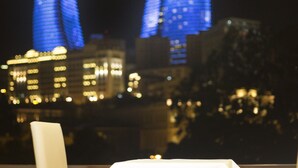 A dining area with a table and chair, a window with a view of illuminated buildings at night. - Old City Inn Hotel (Baku)