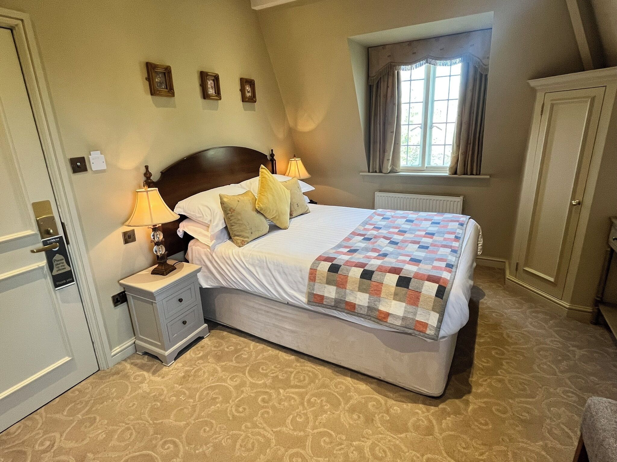 A neatly arranged bedroom with a large bed, bedside lamps, a nightstand, a window with curtains, and a door.