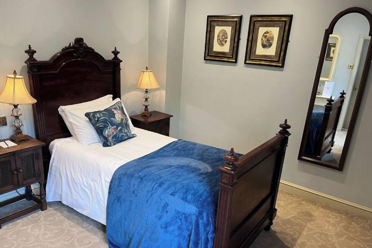 A neatly made bed with a blue blanket, two bedside lamps, and a wooden headboard. There are two framed pictures on the wall and a mirror.