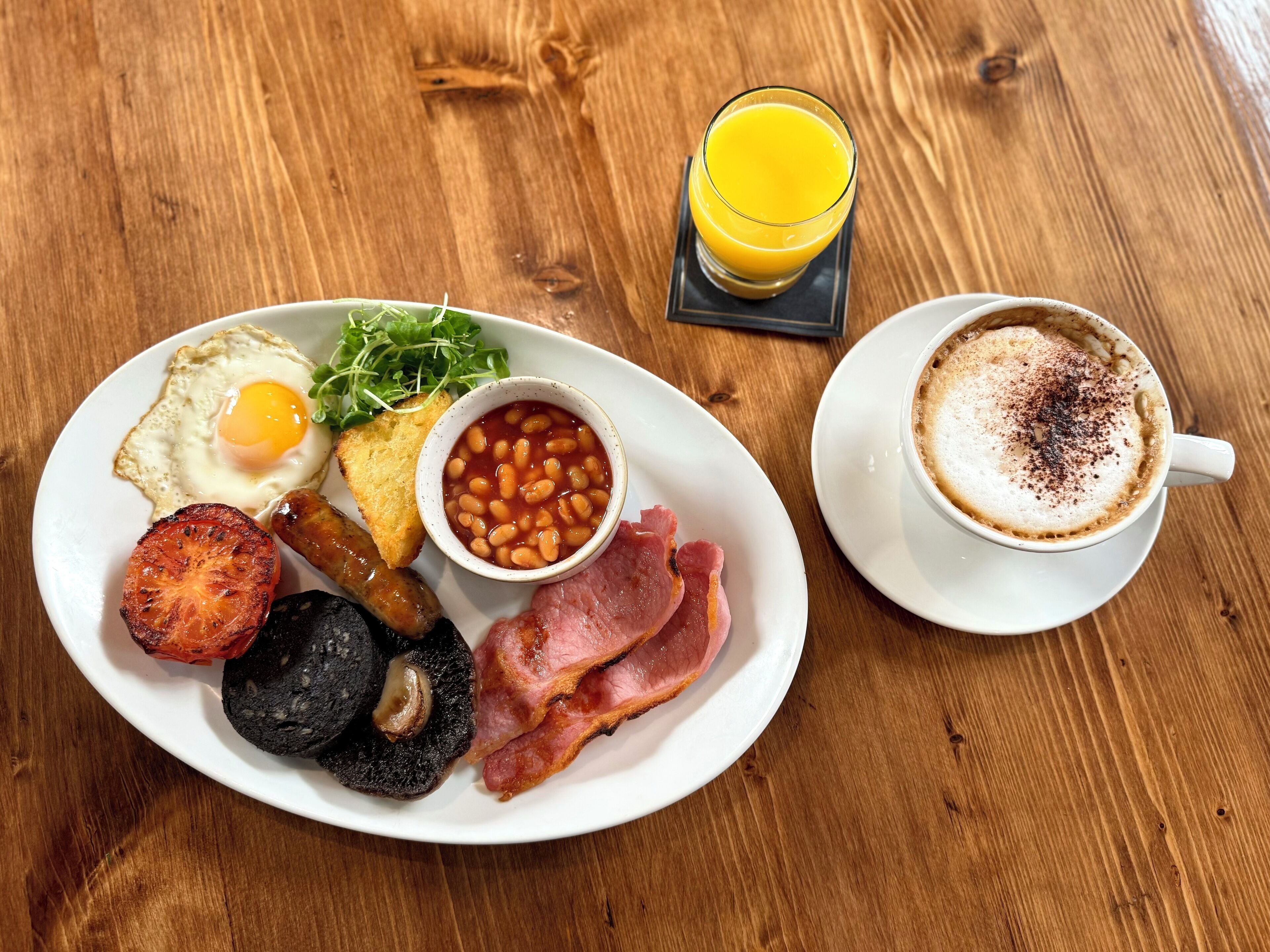Un plato de desayuno con huevos, frijoles, tocino, champiñones y tostadas, junto con un vaso de jugo de naranja y una taza de café.