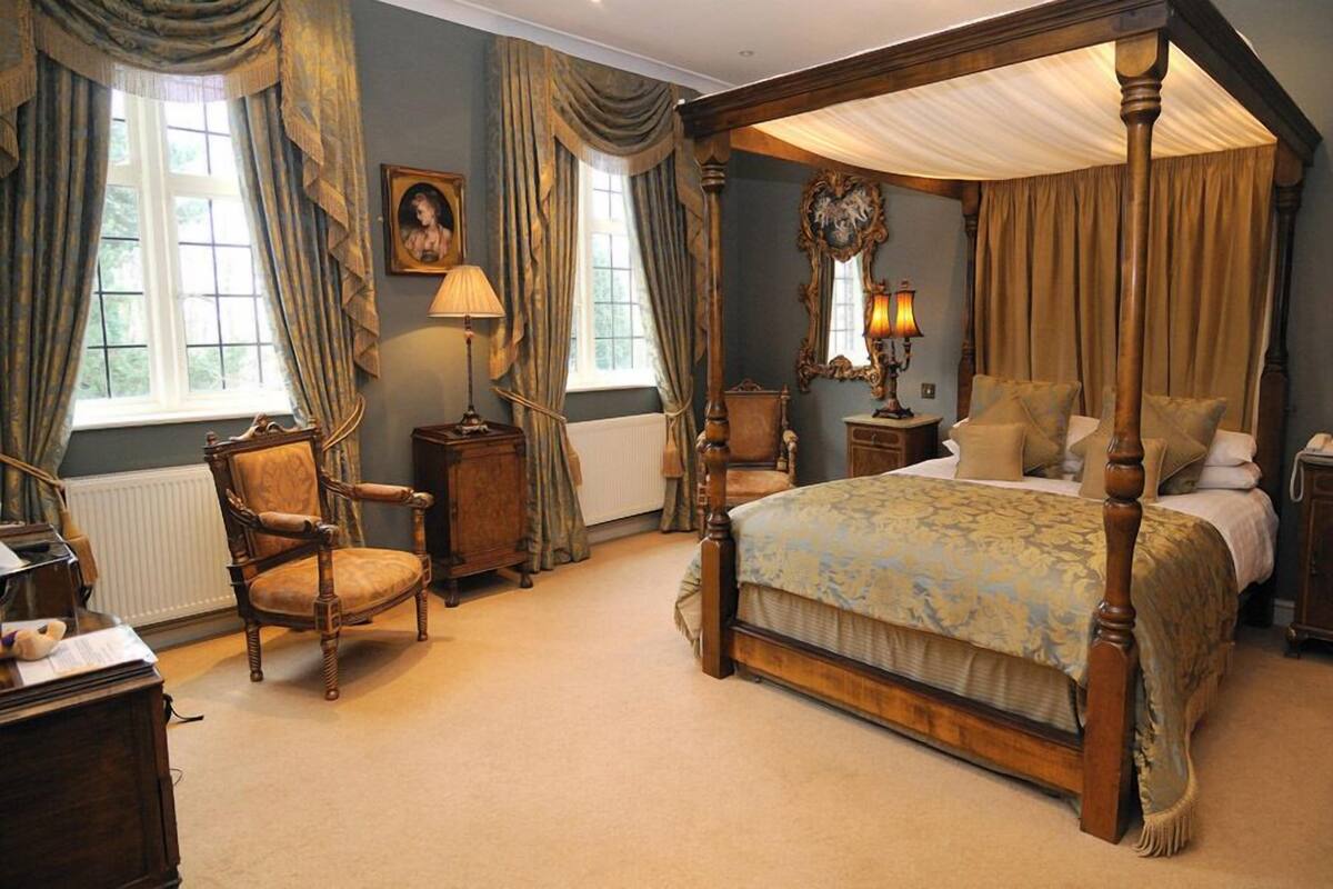 A four-poster bed with a canopy, a wooden chair, a desk with a lamp, and a picture on the wall.