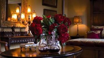 A room with a round table, a vase of red flowers, a bed with red pillows, and a mirror with ornate frames.
