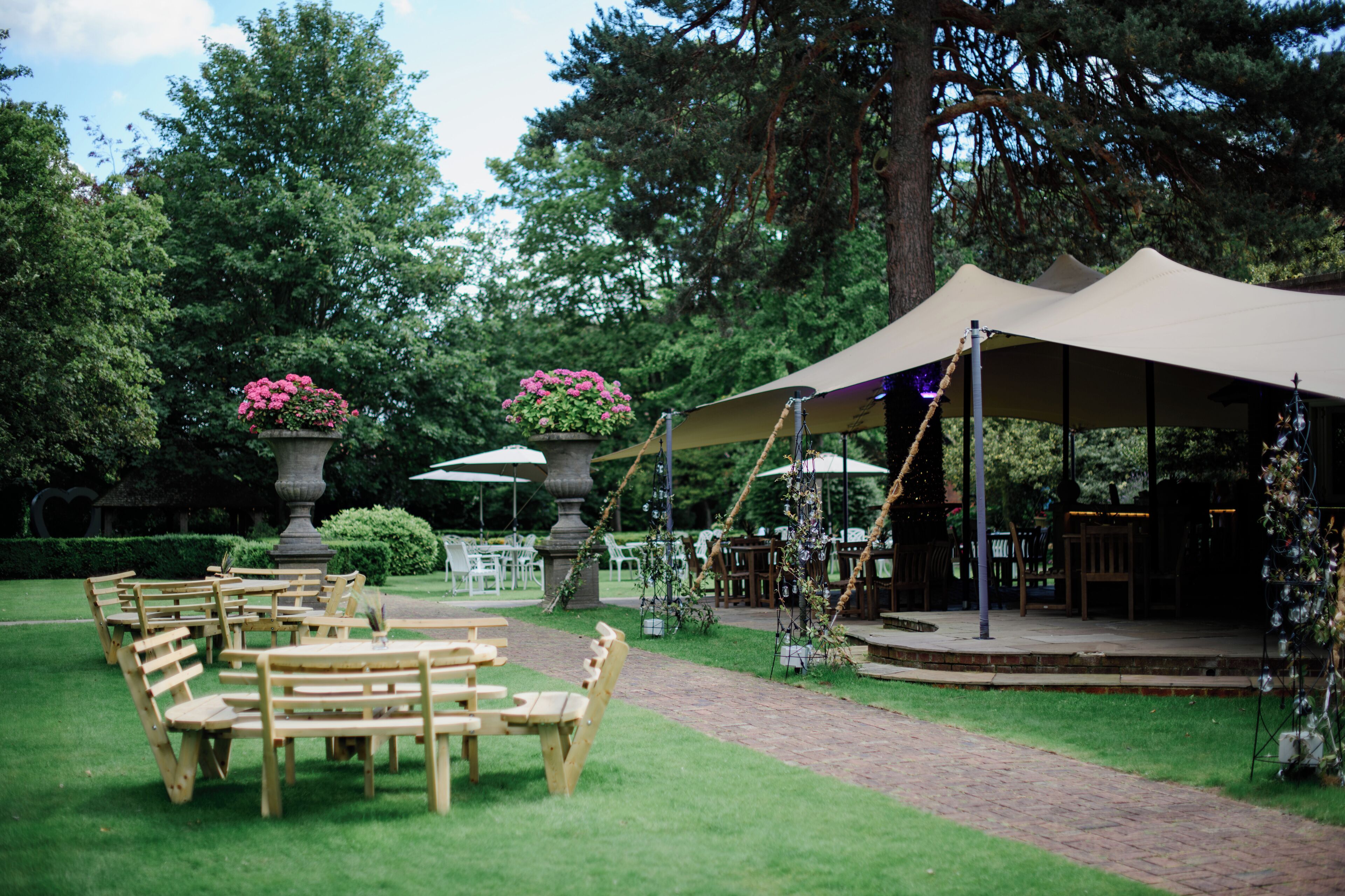 An outdoor seating area with wooden tables and chairs, surrounded by greenery and a covered stage.