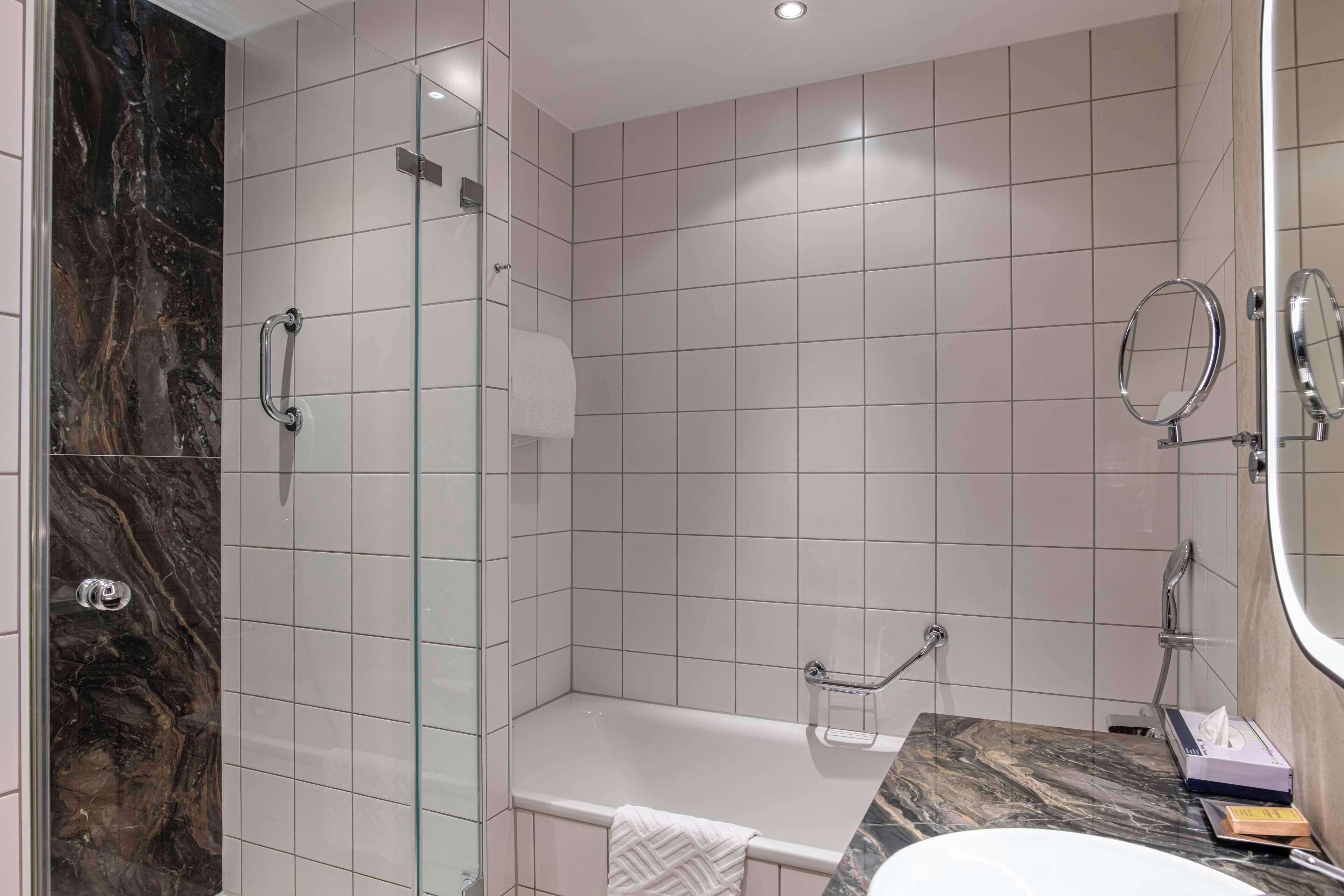 A modern bathroom with a glass shower enclosure, a marble-tiled wall, a white bathtub, and a vanity with a mirror.