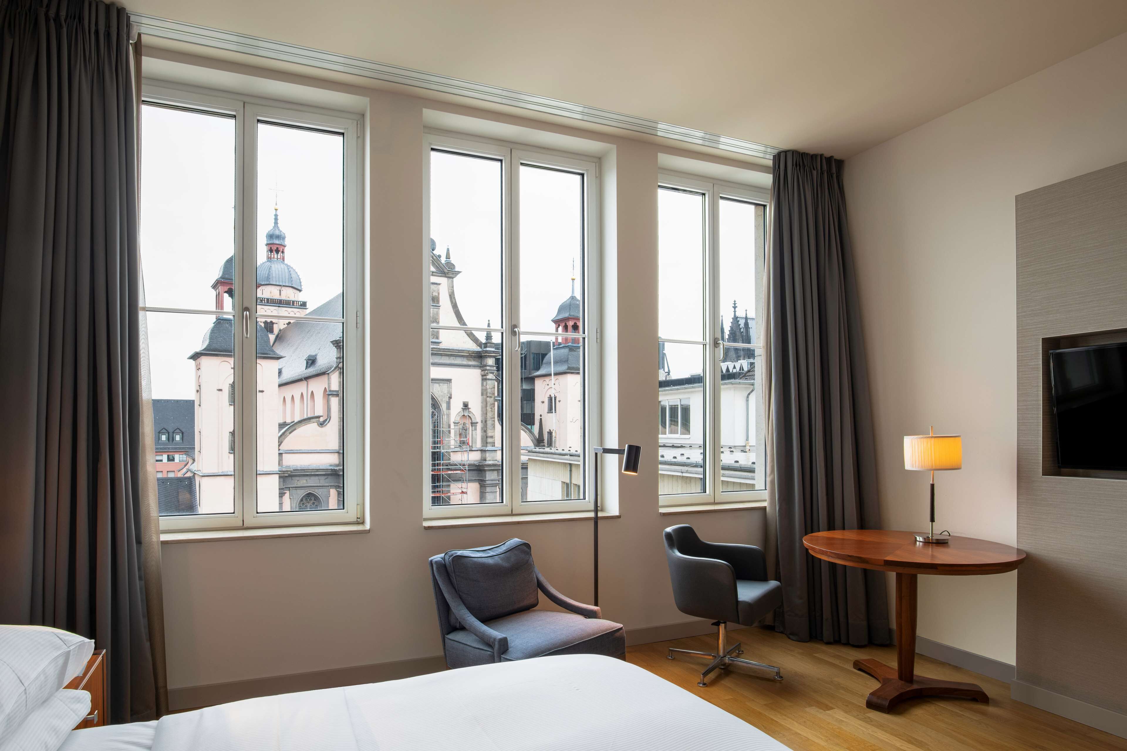 Habitación de hotel con cama, escritorio, silla y ventanas amplias con vista a la ciudad.