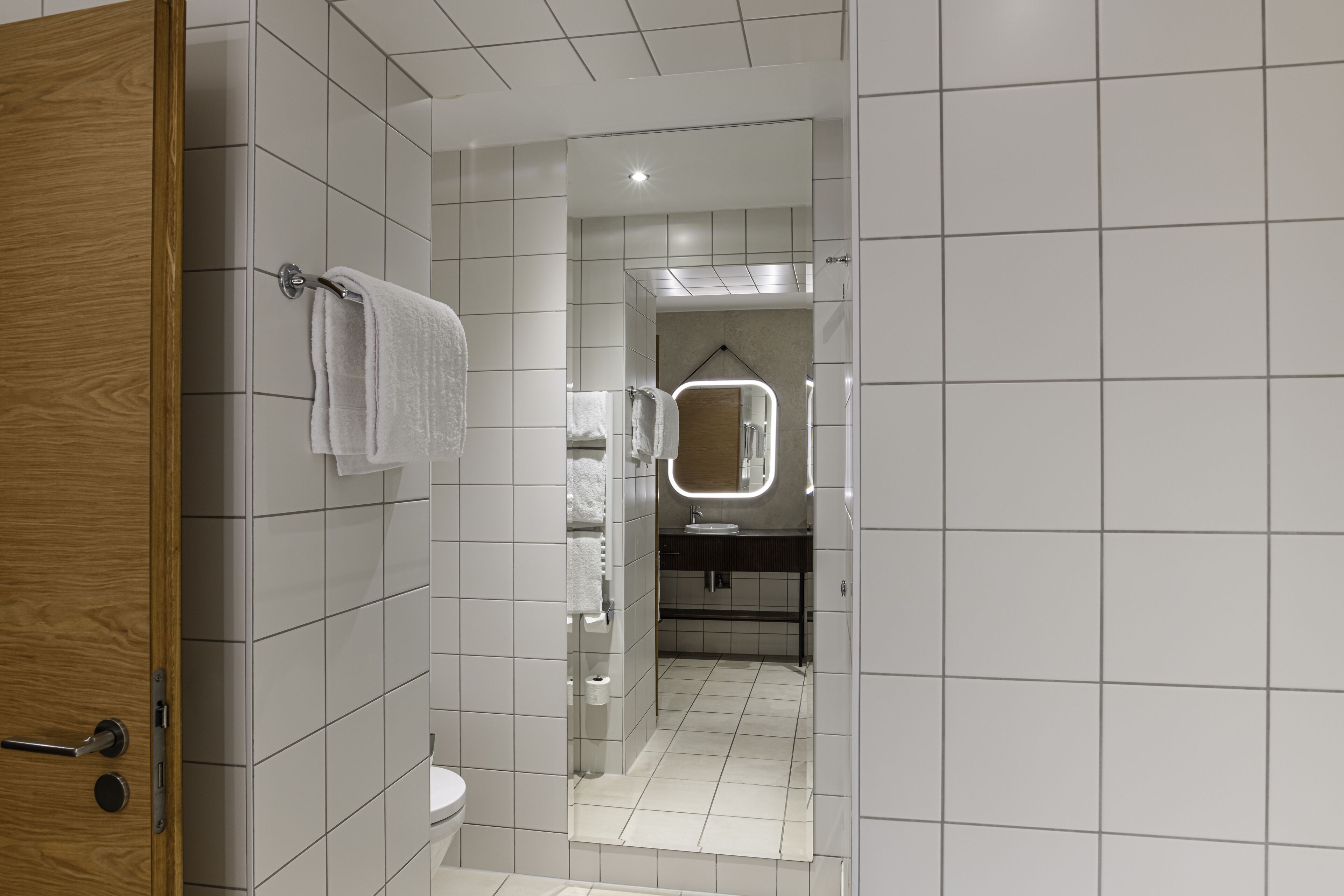 A modern bathroom with white tiles, a wooden door, a mirror with lights, and a shower area.