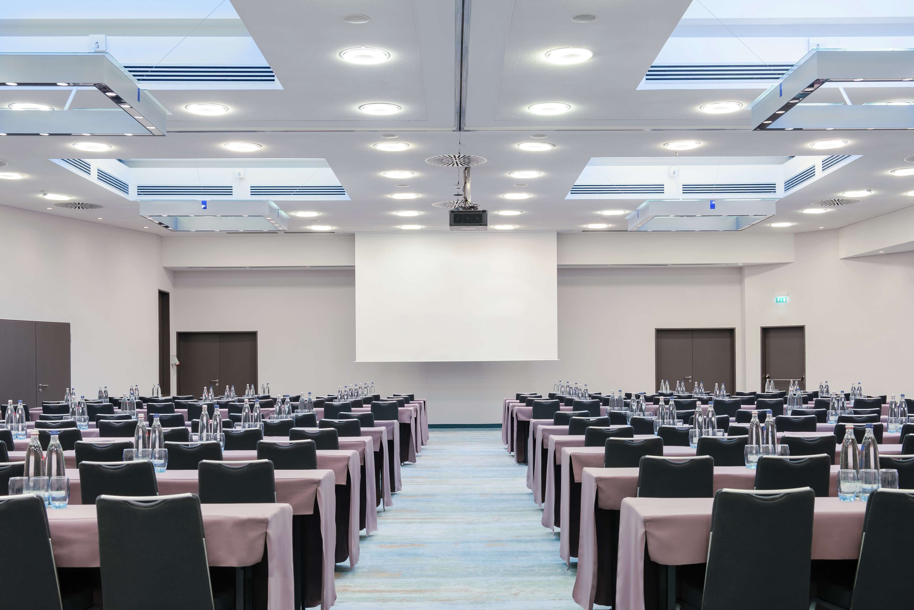A conference room with rows of tables, chairs, and a large screen at the front.