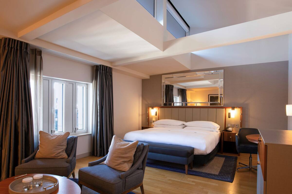 Una habitación de hotel moderna con una cama grande, dos sillones, una mesita con una bandeja y un espejo sobre la cama.
