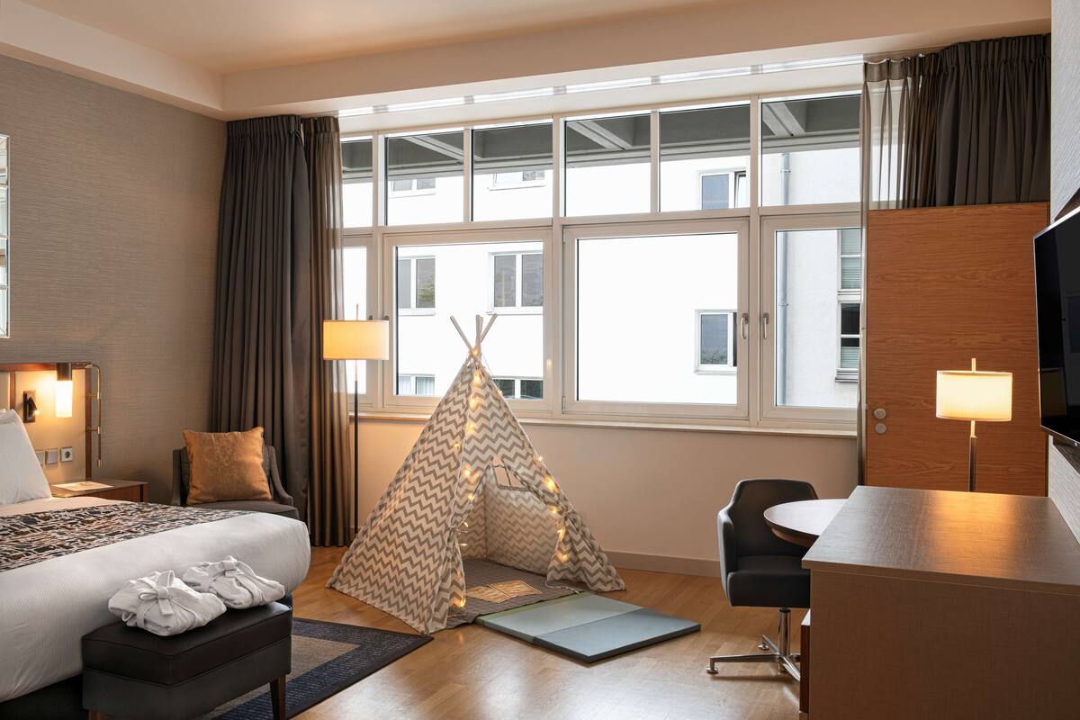 Habitación de hotel con cama, escritorio, silla, una pequeña tienda de campaña y un ventanal grande.