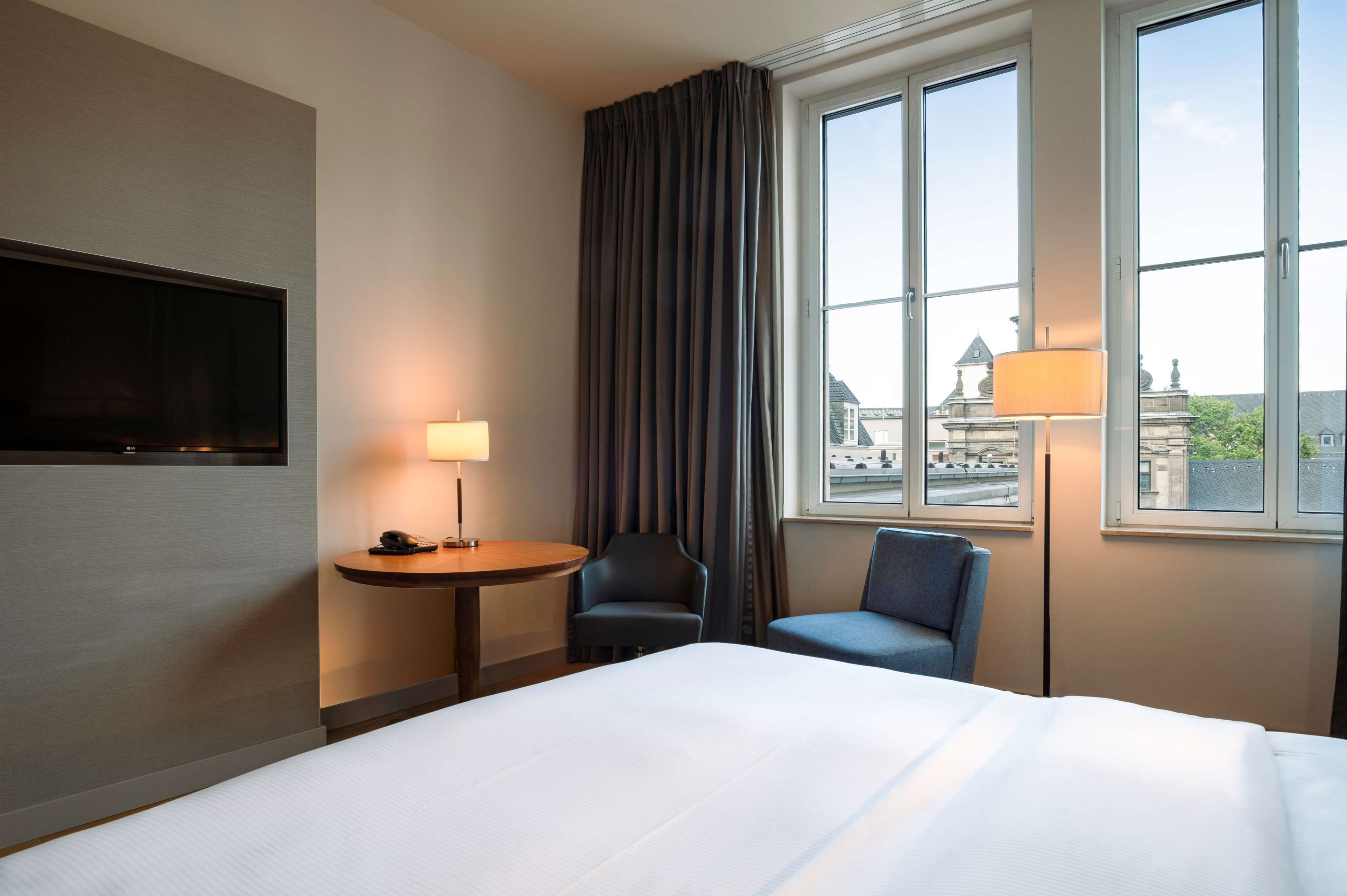 A hotel room with a bed, a desk with a lamp, two chairs, a TV mounted on the wall, and a window with a view of buildings.