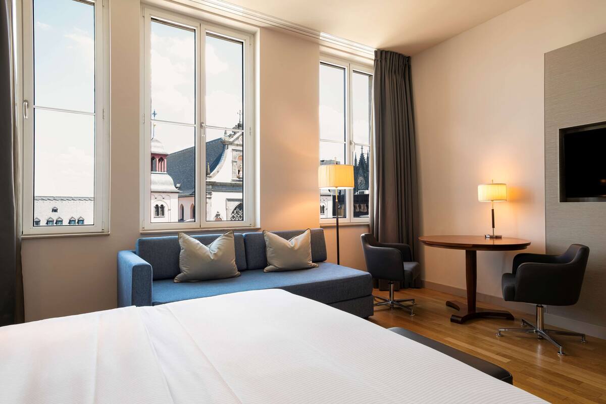 Una habitación de hotel con una cama grande, un sofá azul con almohadas, una mesita redonda con una lámpara y un televisor en la pared.